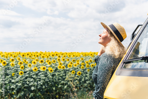 Young blonde woman travelling by campervan. Overlooking sunflower field on sunny summer day photo