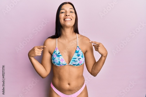 Young latin woman wearing bikini looking confident with smile on face, pointing oneself with fingers proud and happy.