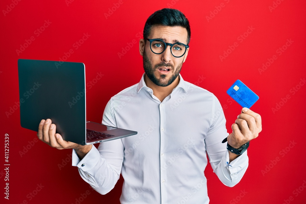 Young hispanic man wearing business style holding laptop and credit card clueless and confused expression. doubt concept. Young hispanic man wearing business style holding laptop and credit card clueless and confused expression. doubt concept.