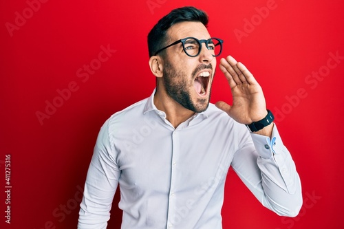 Young hispanic businessman wearing shirt and glasses shouting and screaming loud to side with hand on mouth. communication concept. © Krakenimages.com