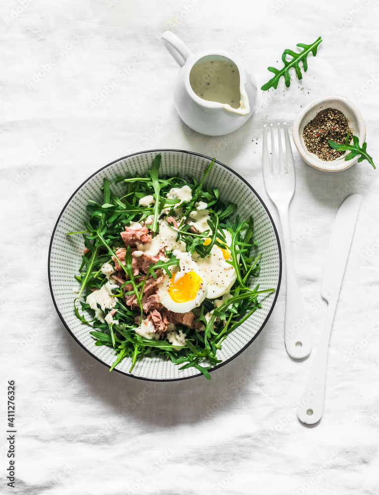 Salad with arugula, canned tuna, boiled egg and yogurt, mustard dressing on a light background, top view