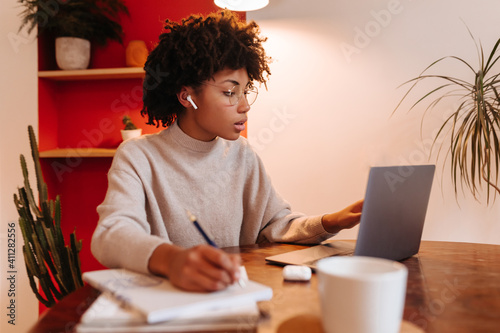 Dark-skinned girl in glasses and headphones looks at laptop screen and writes notes in notebook