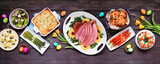 Classic Easter ham dinner. Top view table scene on a dark wood banner background. Ham, scalloped potatoes, eggs, hot cross buns, carrot cake and vegetables.
