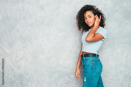 Beautiful black woman with afro curls hairstyle.Smiling model in white trendy jeans clothes. Sexy carefree female posing near gray wall in studio. Tanned and cheerful