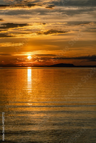 Vertical photo of Sunset over Umea river Mountains, summer sky with clouds highlighted by orange red Sun. Sunlight path on water Storuman, Lapland, Sweden