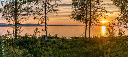 Scenic Panorama - Sunset over Umea river Mountains, summer sky with clouds highlighted by orange red Sun. Blurry foreground with trees at coastline. Sunlight path on water Storuman, Lapland, Sweden photo