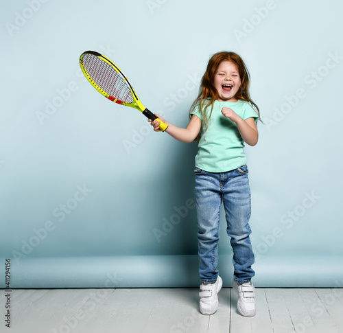 Beautiful red-haired girl playing tennis isolated studio shot. Little baby-player in a trendy outfit ready to hit the ball with the racket and screaming loudly. Children's sports, hobby for an active © FAB.1