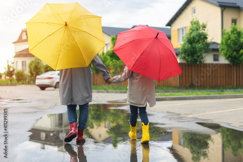 Mom and daughter rainy weather go home under umbrellas and in rubber boots. mother and child walk down the street in autumn through puddles