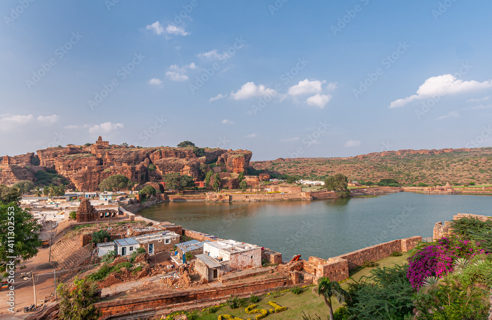 Badami, Karnataka, India - November 7, 2013: Agasthya Lake and surrounding red rock hills. Some brown stone temple buildings sprinkled around. Green foliage and pink flowers add color.