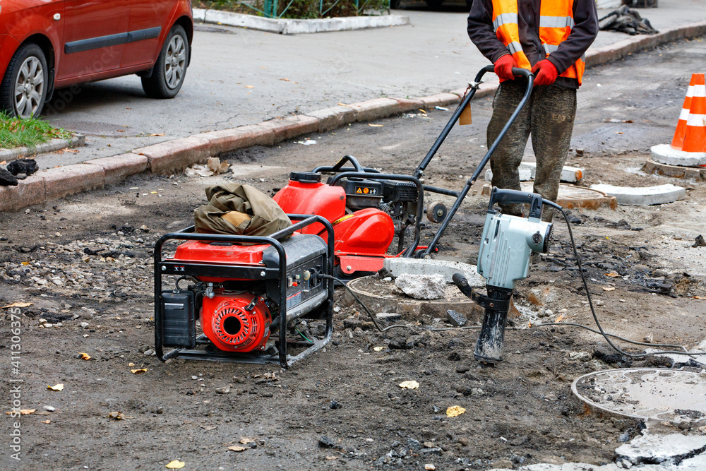 A road worker repairs and installs sewers on the road using a compactor plate, an electric jackhammer and a petrol generator.