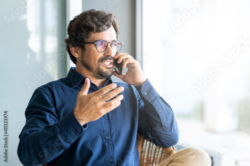 Casual businessman wearing glasses talking on the phone. Successful male portrait sitting on a shair at the office by the window