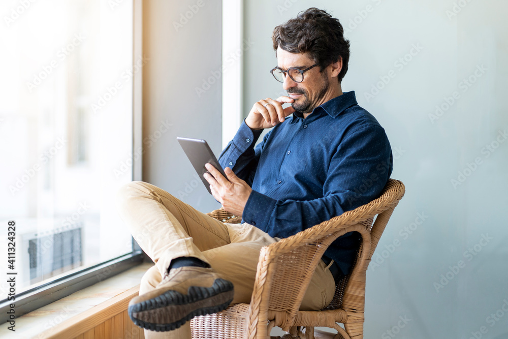 Casual business man wearing glasses sitting looking at his tablet. Successful male portrait with thinking face expression working from home by the window Casual business man wearing glasses sitting looking at his tablet. Successful male portrait with thinking face expression working from home by the window