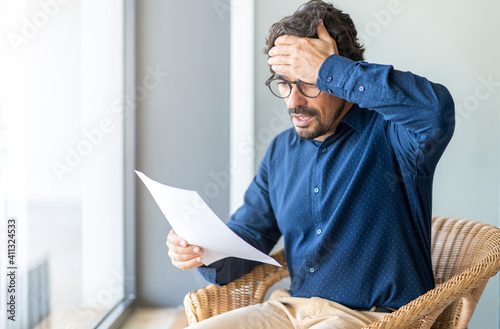 Casual man looking surprised reading bills to pay. Taxes, bank statement and loan debt. Male portrait with worried face and hand on his head