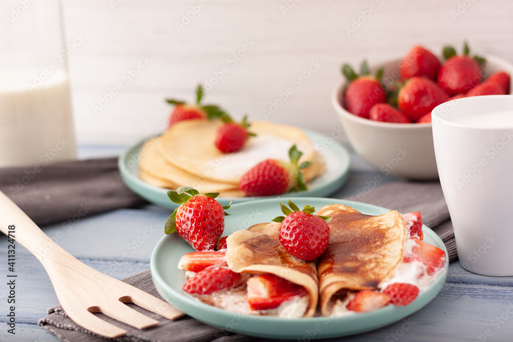 Imagen de un plato de crepes con trozos de fresas y nata y otro detras con azucar glass sobre un fondo claro y un cuenco de fresas con una taza y una botella de leche. Imagen de un plato de crepes con trozos de fresas y nata y otro detras con azucar glass sobre un fondo claro y un cuenco de fresas con una taza y una botella de leche.