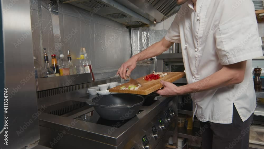 Slow-motion midsection shot of unrecognizable male cook in white jacket adding fresh vegetables from cutting board to wok pan for frying Slow-motion midsection shot of unrecognizable male cook in white jacket adding fresh vegetables from cutting board to wok pan for frying