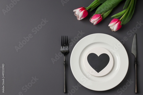 Creative table setting with hearts on a white plate, black fork and knife, tulips on a dark background. Valentine's Day, Wedding Day, Birthday, Women's Day and Mother's Day. Flat lay. Copy space