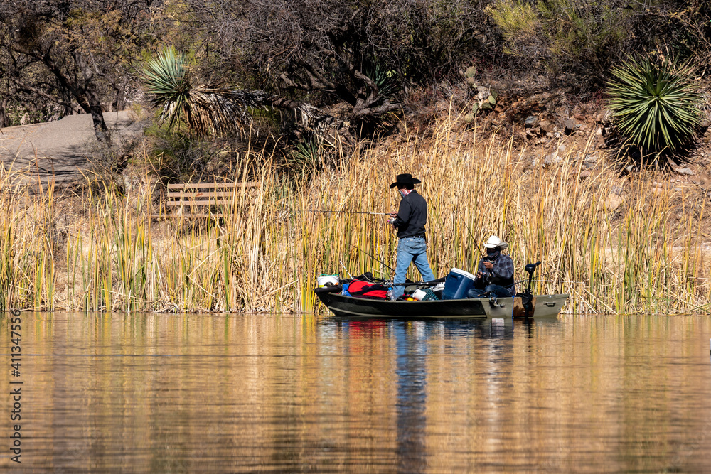 Two people fish from a small boat at Patagonia Lake State Park, Arizona. Both men are wearing face coverings. Their boat is overfilled with gear. Concepts of outdoor recreation, fishing, state parks