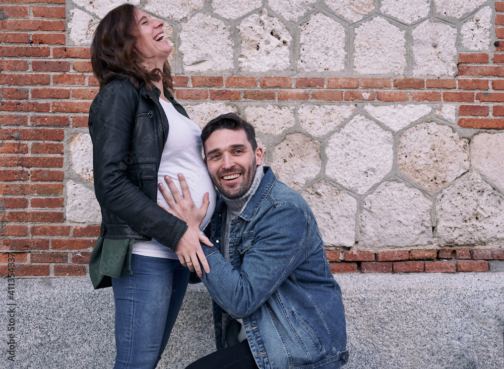 A pregnant woman standing in front of a brick building. Man hugs her belly. Couple.