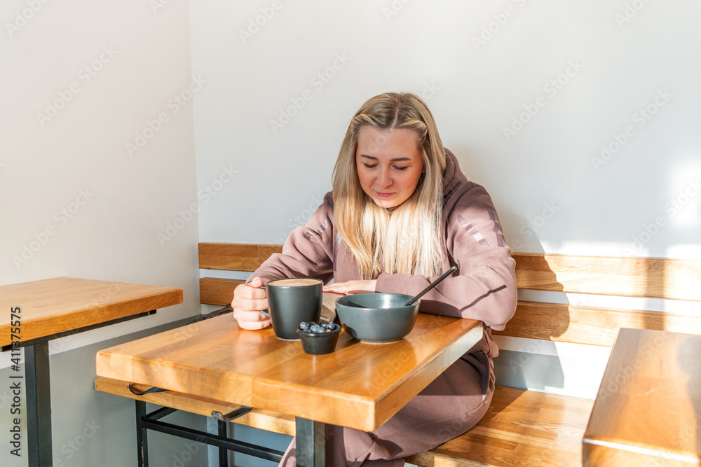 Young woman in a comfortable tracksuit is reading a paper book while drinking coffee in a cafe. Young woman in a comfortable tracksuit is reading a paper book while drinking coffee in a cafe.