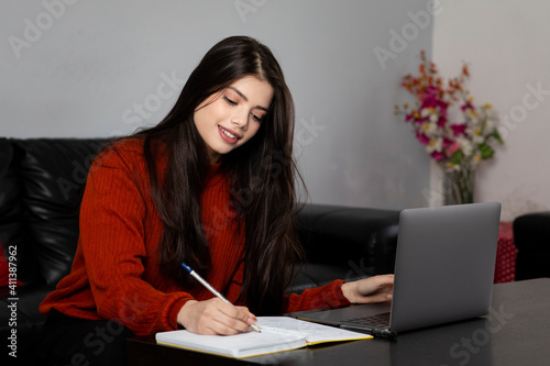 A young Indian girl writes notes in her diary while studying from home from her laptop.