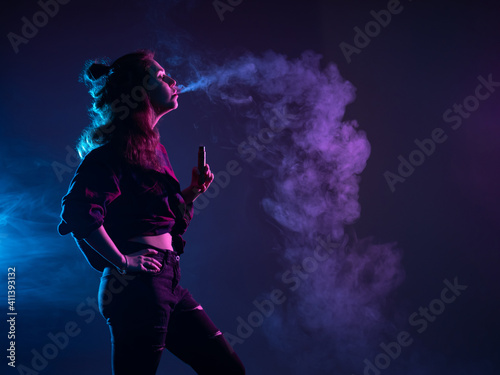 A girl with a VAPE stands in profile. A woman smokes a VAPE and lets out smoke. Girl vaper on a dark background. A girl with an e-cigarette looks at the smoke. Vaping concept with place for text. © Grispb