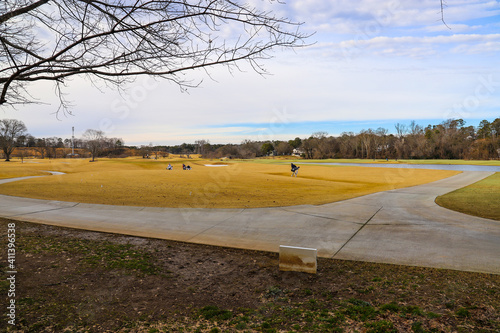 A majestic shot of a golf course with autumn colored grass surrounded by green and autumn colored trees with blue sky and clouds with people walking along the course playing golf at Tanyard Creek Park photo