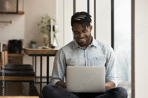 Skilled african man freelancer sit in comfortable pose work from home consult client by video link write programme code on pc. Young black guy relax at home with laptop post comments at social network