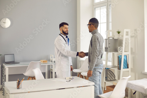 Friendly young male doctor shakes hands with his dark-skinned patient welcoming him to a modern medical clinic. Healthcare, health insurance, partnership, trust and medical ethics concept.