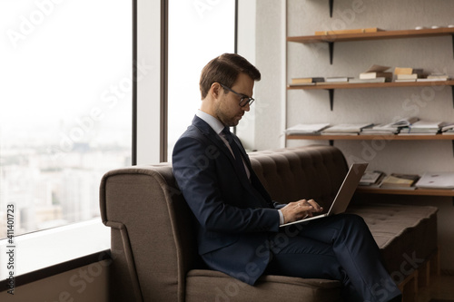 Confident businessman in formal suit and glasses work on laptop hold device on knees while expecting for business meeting. Serious young man visitor sit on sofa in office waiting room using computer