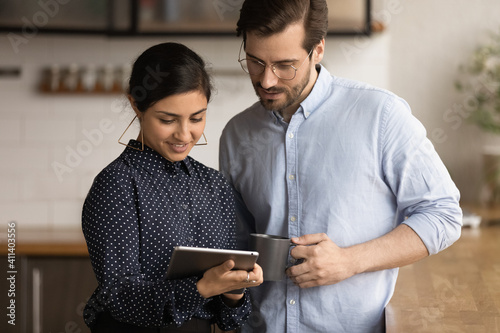 Smiling millennial wife of indian ethnicity sharing good news on digital tablet screen with caucasian husband. Diverse multiethnic colleagues reading document using pad at office kitchen at break time