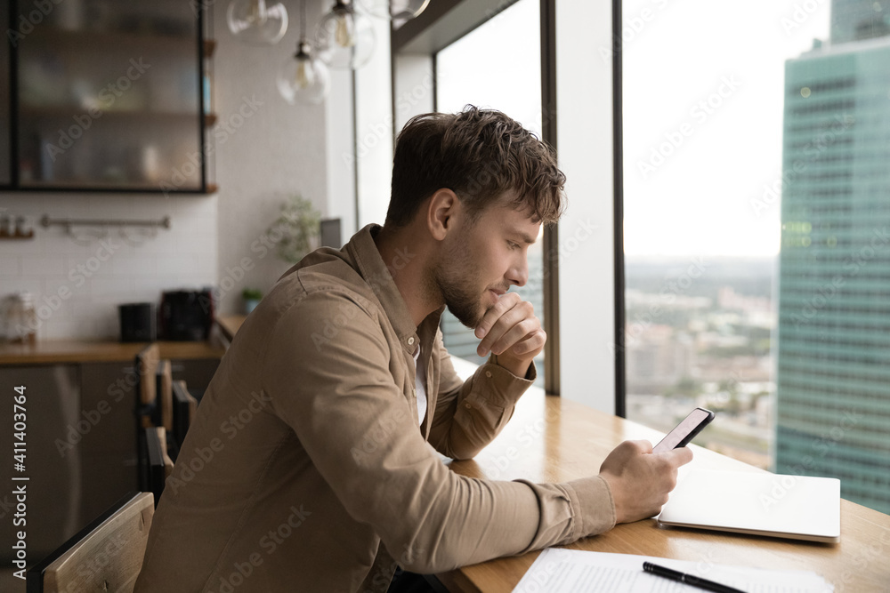 Fototapeta premium Information to think about. Pensive male freelancer look at cell ponder on job offer received by email. Young businessman sit by large window at modern kitchen read important message on phone screen