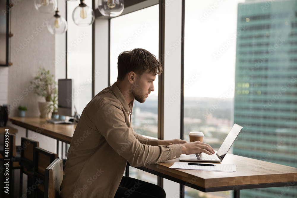 Concentrated millennial man write message on laptop keyboard at desk near panoramic window with splendid cityscape. Side shot of attentive young guy working on project document at modern office online