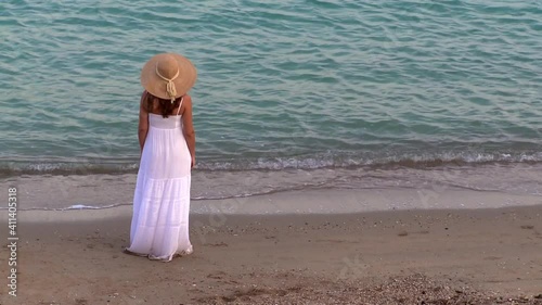 Woman alone in a white dress and hat standing in front of the sea on a deserted beach thinking, nostalgia, emotions, memories 