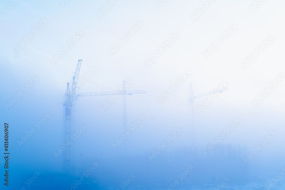 Tall tower cranes shine through in the winter morning fog. Bird's-eye view of the construction site on a foggy morning.