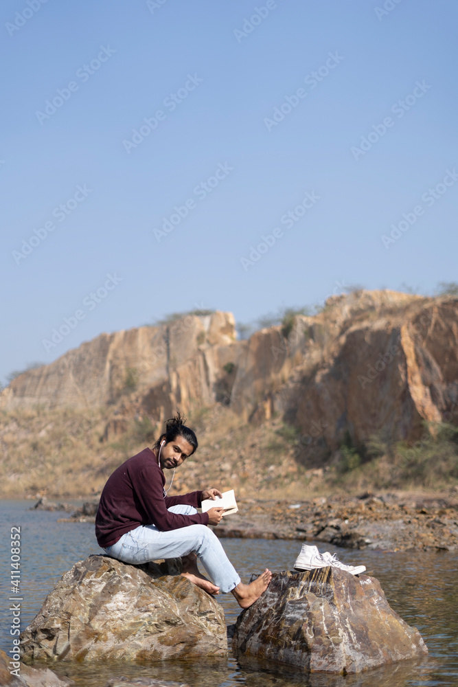 Young indian traveler with long hair sitting near a lake with some books. travel and freedom concept.