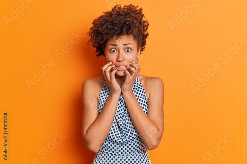 Nervous worried Afro American woman clenches teeth looks embarrassed at camera wears fashiobnable outfit afraids of bad consquences poses in studio against orange background. Scared female model photo
