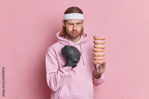 Serious displeased European male boxer makes fist punch ready for fight wears sportclothes holds doughnuts dressed in sweatshirt isolated over pink background. Be strong and healthy concept. photo