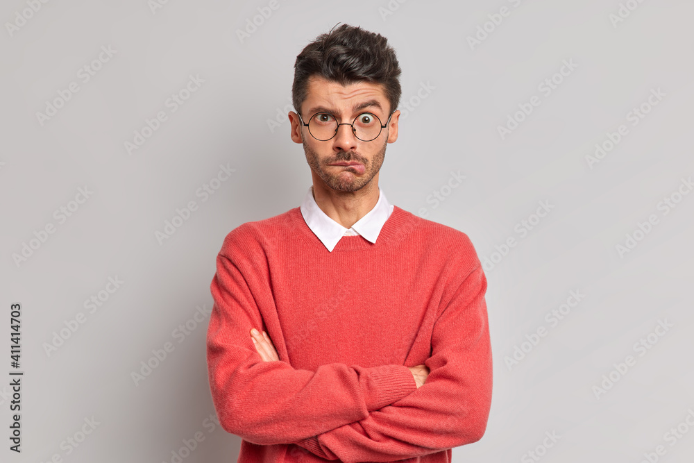 Displeased Caucasian man bites lips keeps hands crossed dissatisfied with something wears round spectacles and sweater looks offended at camera poses against grey background. Negative emotions Displeased Caucasian man bites lips keeps hands crossed dissatisfied with something wears round spectacles and sweater looks offended at camera poses against grey background. Negative emotions