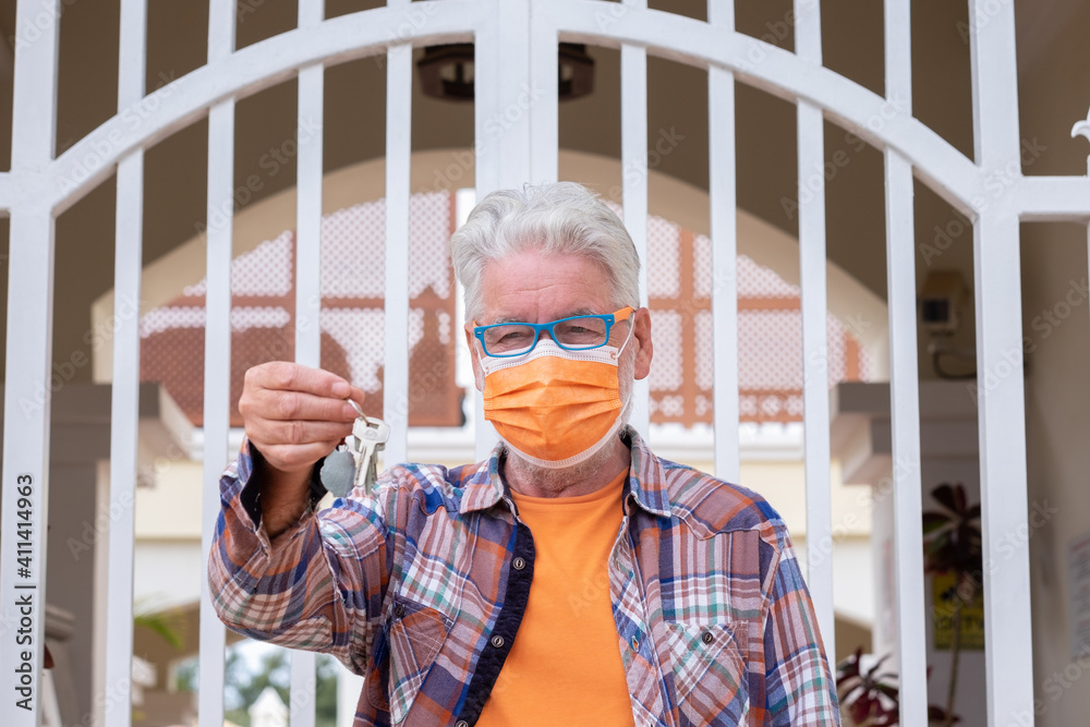 Coronavirus. Senior man wearing a surgical mask due to coronavirus standing behind his home's gate. An elderly man with white hair holding home keys Coronavirus. Senior man wearing a surgical mask due to coronavirus standing behind his home's gate. An elderly man with white hair holding home keys