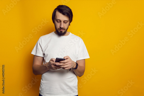 man with a beard and a white T-shirt stands with a phone on a yellow background © saulich84