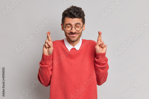 Fotografia Positive happy man closes eyes believes dreams come true hopes to get promotion at work crosses fingers dressed in casual red jumper isolated over grey background