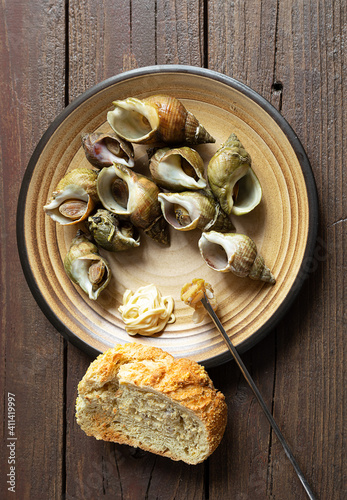 Fresh sea snails bulot or common whelks prepared according to a traditional French recipe in a plate and a piece of bread on a dark wooden table, top view photo