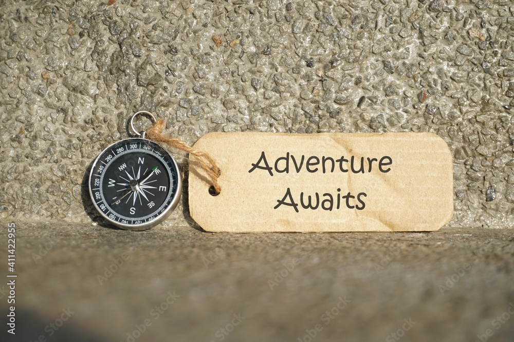 Fototapeta premium Conceptual Image: Magnetic compass with paper tag written Adventure Awaits, selective focus.