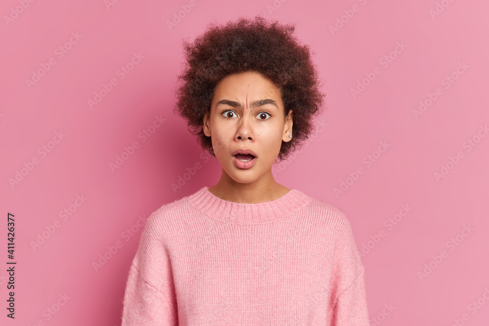 Photo of puzzled surprised African American woman stares at camera with great wonder and disbelief keeps mouth opened dressed in casual jumper isolated over pink background. Face expressions Photo of puzzled surprised African American woman stares at camera with great wonder and disbelief keeps mouth opened dressed in casual jumper isolated over pink background. Face expressions