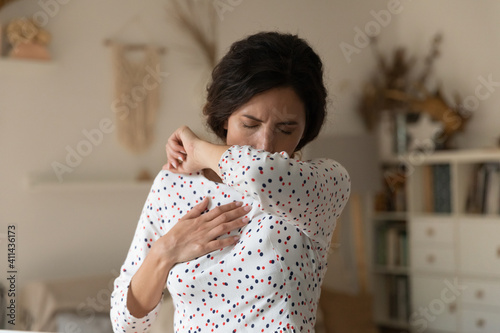 Unhealthy millennial Caucasian woman feel sick cough in elbow suffer from cold or influenza. Unhappy unwell young female struggle with bronchitis or corona virus symptoms at home. Covid-19 concept. photo