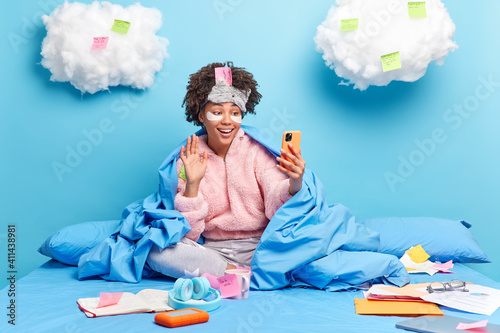 Happy millennial girl with Afro hair waves hello in smartphone device does reserach or assignment at home enjoys domestic atmosphere poses on bed applies patches under eyes makes video call. photo