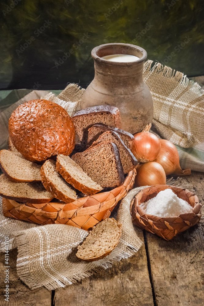 Rye bread on a vintage wooden table in a rustic style is cut into pieces in an old wicker birch bark dish with milk in an old clay pot, salt and onions on a canvas tablecloth