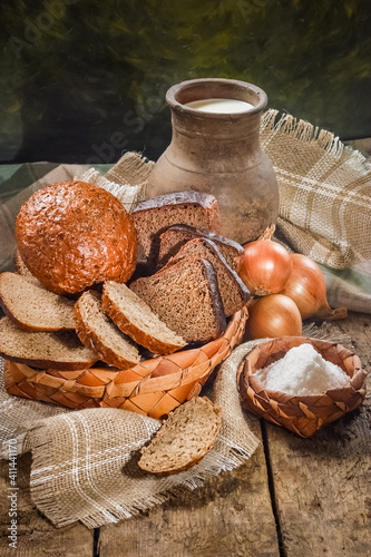 Obraz na płótnie Rye bread on a vintage wooden table in a rustic style is cut into pieces in an old wicker birch bark dish with milk in an old clay pot, salt and onions on a canvas tablecloth w salonie
