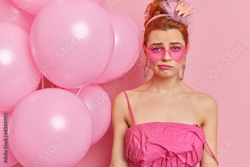 Displeased young female with professional makeup wears pink sunglasses and dress looks unhappily at camera has bad mood on birthday party dissatisfied with something poses near balloons indoor © wayhome.studio 