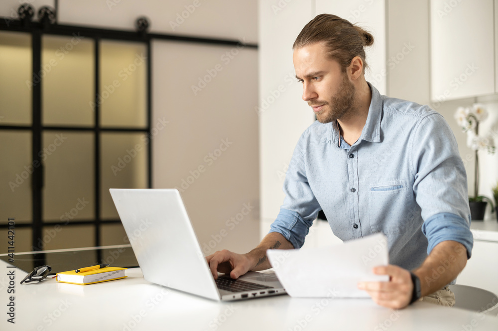 Concentrated guy with a laptop is working on the project remotely, young entrepreneur looks through the work papers, analyzing, checking datas. A male office employee prepares a report Concentrated guy with a laptop is working on the project remotely, young entrepreneur looks through the work papers, analyzing, checking datas. A male office employee prepares a report